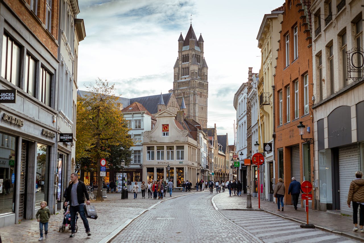 Home Picturesque street in Bruges with Belfry tower, cobblestones, and people strolling in autumn.